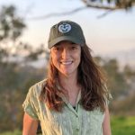 Georgia Titcomb standing amidst nature, wearing a CSU Rams baseball cap and smiling at the camera. Behind a pastel sunset can be seen in the distance.