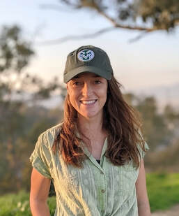 Georgia Titcomb standing amidst nature, wearing a CSU Rams baseball cap and smiling at the camera. Behind a pastel sunset can be seen in the distance.