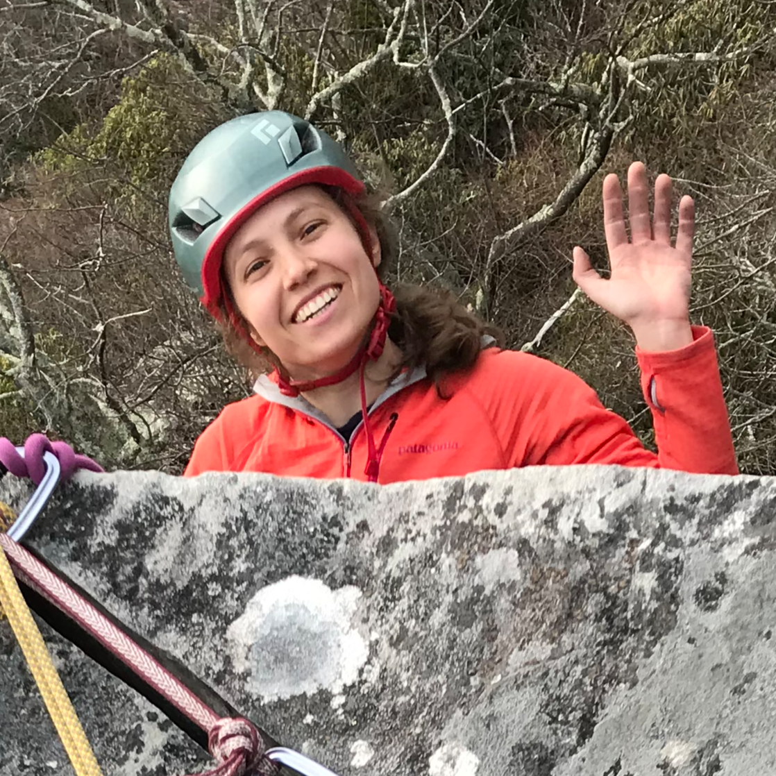 Andee Kaplan waving at the camera while peeking up over a rock while rock climbing. She is in full gear with helmet, ropes, and cables.