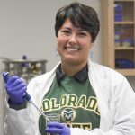 Woman in white lab coat and CSU sweatshirt working with lab equipment and smiling at the camera