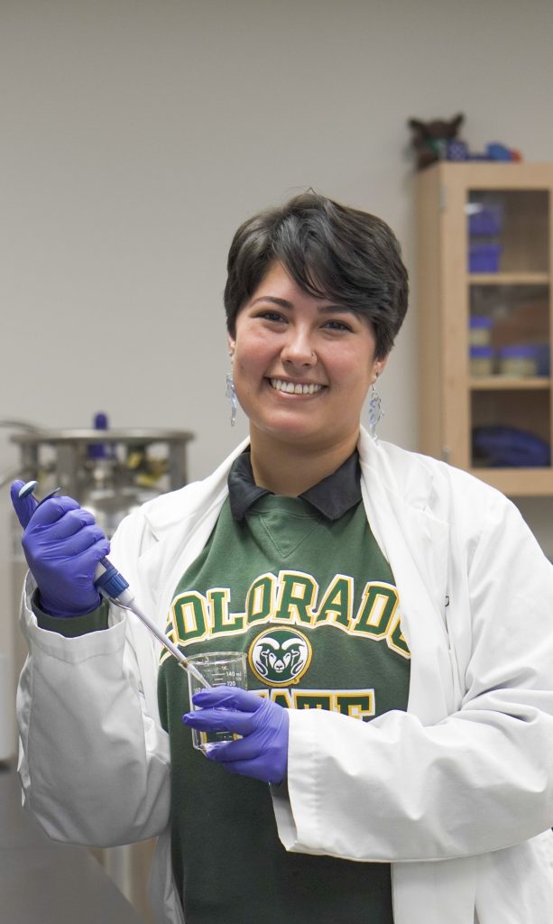 Woman in white lab coat and CSU sweatshirt working with lab equipment and smiling at the camera
