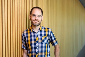 Dr. Devin O'Connor in a black, yellow, and white plaid shirt in front of a wall of vertical wooden slats extending down a long hallway.