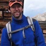James Adelman in backpack, hat, and glasses, with an old wooden structure in the background and mountains beneath a cloudy sky can be seen through the window frame.
