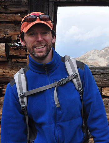James Adelman in backpack, hat, and glasses, with an old wooden structure in the background and mountains beneath a cloudy sky can be seen through the window frame.