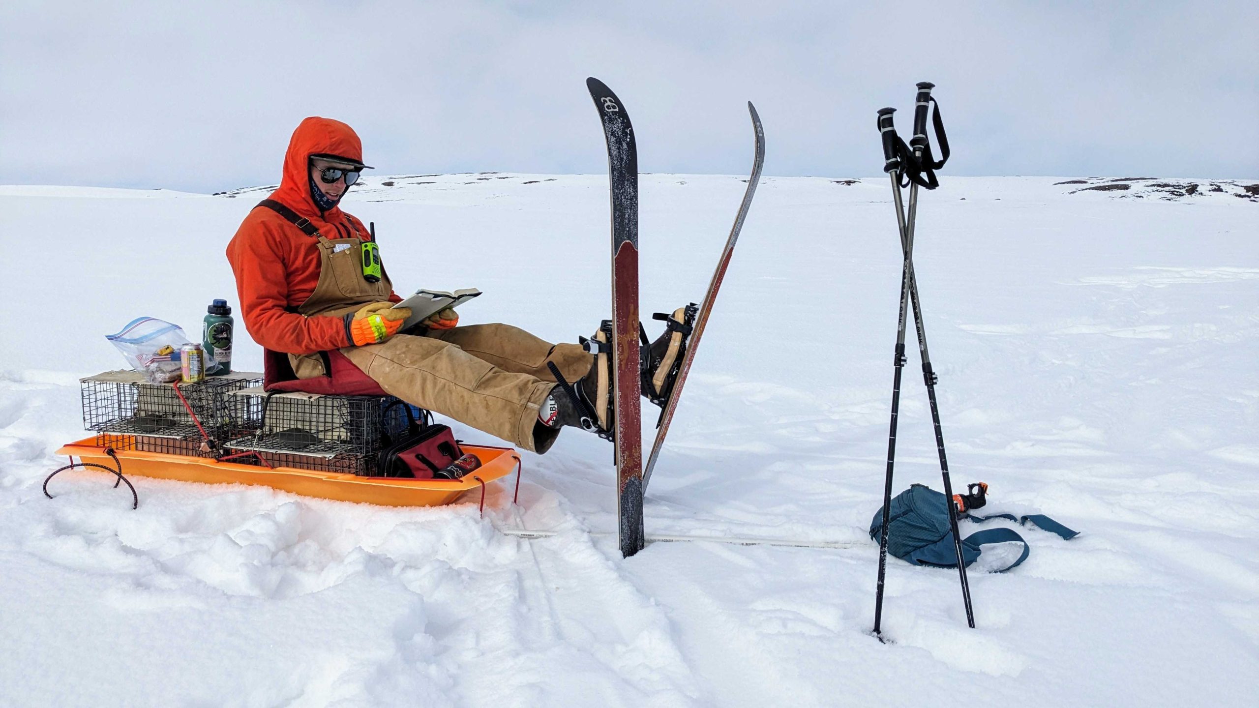 Cole Deal wearing an orange jacket and tan overalls sitting on a sled in a snowy landscape, reading a book. His skis are propped upright in the snow nearby, along with ski poles and a backpack. Field equipment, including a metal live trap and water bottle, is secured on the sled beside them. The scene is set on an open, snow-covered expanse under an overcast sky.