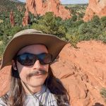 Andrew Paton wearing a wide‑brimmed sun hat, sunglasses, and a light T‑shirt stands on red rock terrain overlooking the dramatic sandstone formations and green valleys of Garden of the Gods on a sunny day.