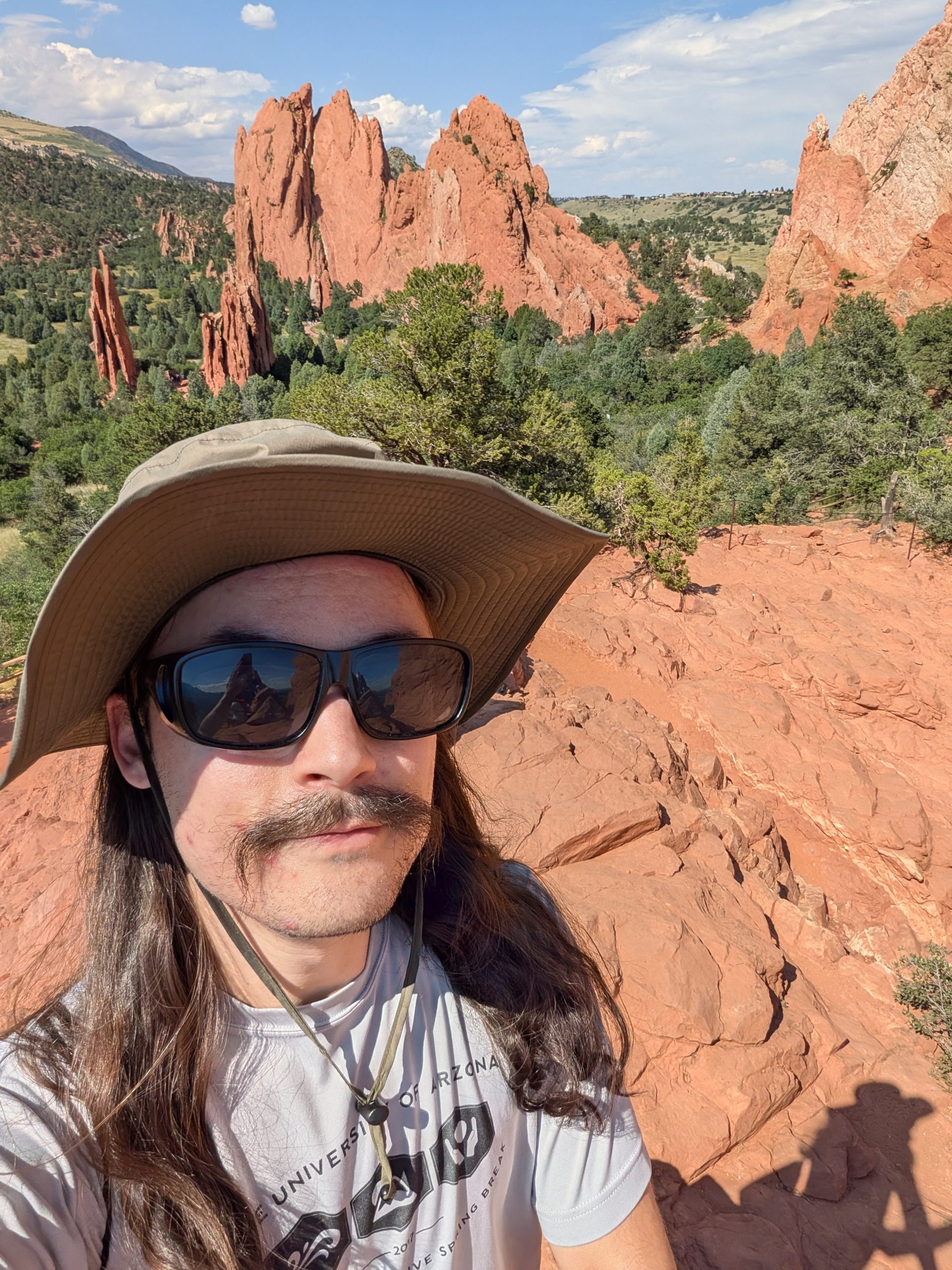 Andrew Paton wearing a wide‑brimmed sun hat, sunglasses, and a light T‑shirt stands on red rock terrain overlooking the dramatic sandstone formations and green valleys of Garden of the Gods on a sunny day.