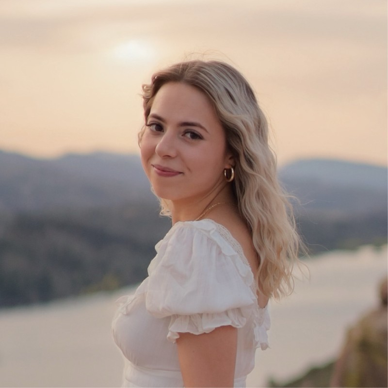 Paige standing at a three quarter profile looking over her shoulder and looking back at the camera with a faint smile. A Colorado sunset over Horsetooth Reservoir can be seen in the background in pale pinks, oranges, and purples.