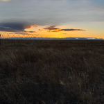 A scenic image of a grassy field at sunset with vibrant orange and yellow hues lighting up the sky. A person is silhouetted against the horizon, standing near a series of curved metal structures resembling a greenhouse frame. Mountains can be seen in the distance under a dramatic sky with scattered clouds. The peaceful landscape evokes a serene and tranquil atmosphere.