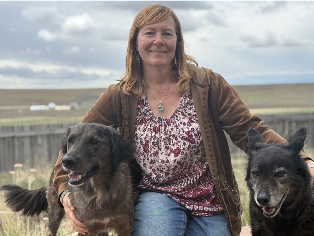 Robyn Bortner posing with two dogs on either side of her. She's outside beneath an overcast cloudy sky with a wide open prairie and ranch land behind her.