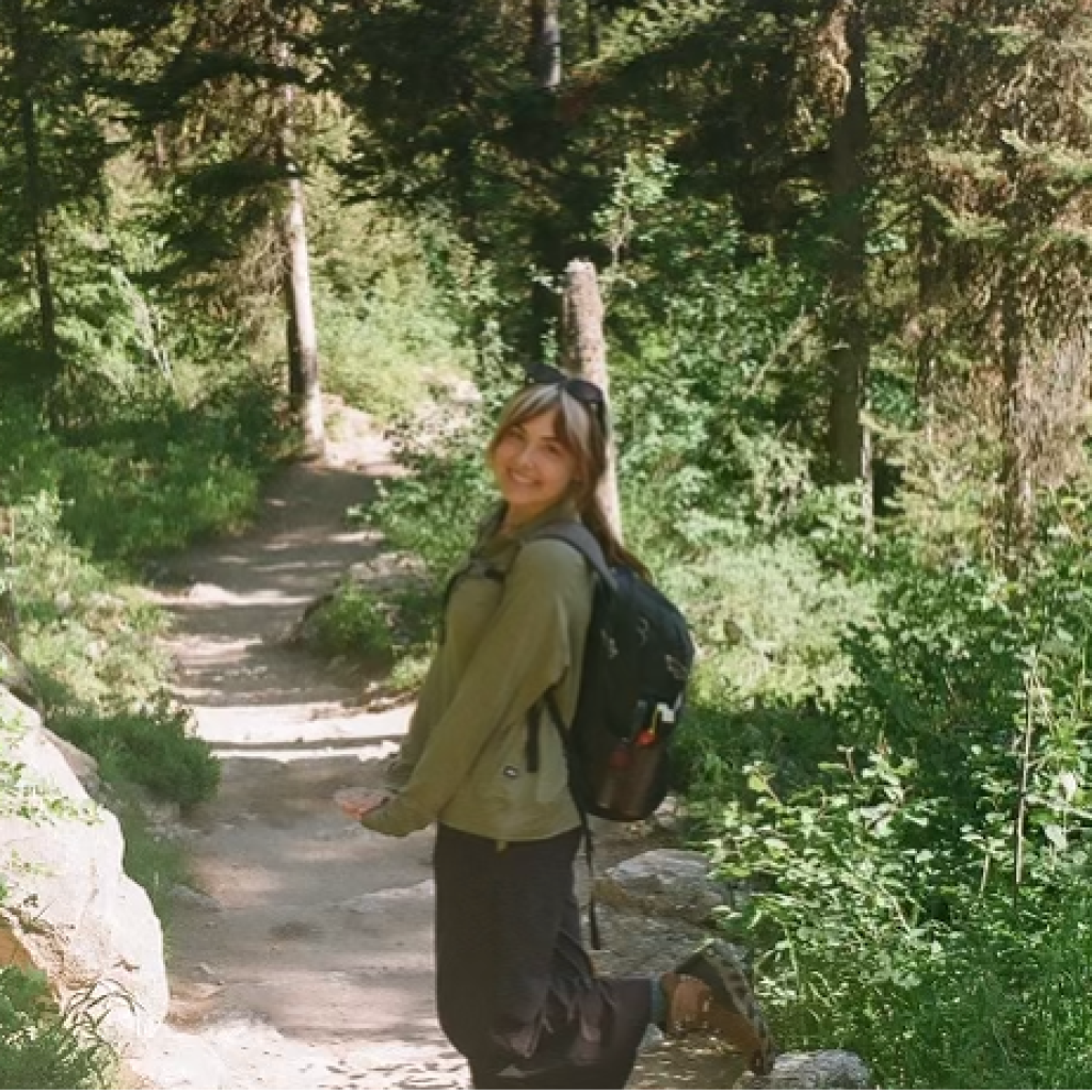 Anna Hall stands on a narrow dirt trail surrounded by lush green vegetation and tall trees in a forested area. She is wearing a light green jacket, dark pants, and sandals, with a black hiking backpack and sunglasses. The sunlight filters through the trees, creating a serene and natural atmosphere.