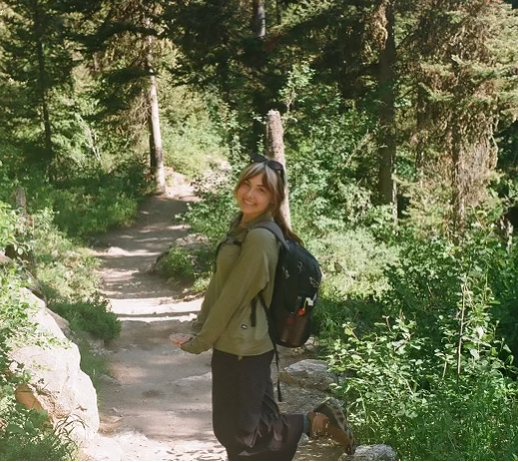 Anna Hall stands on a narrow dirt trail surrounded by lush green vegetation and tall trees in a forested area. She is wearing a light green jacket, dark pants, and sandals, with a black hiking backpack and sunglasses. The sunlight filters through the trees, creating a serene and natural atmosphere.