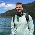 Scott Otto stands outdoors on a dock wearing a light green patterned hoodie, sunglasses hanging from the neck of his shirt, and a black backpack. Behind him is a scenic view of a lake surrounded by lush green forest and mountains under a bright blue sky with scattered clouds.
