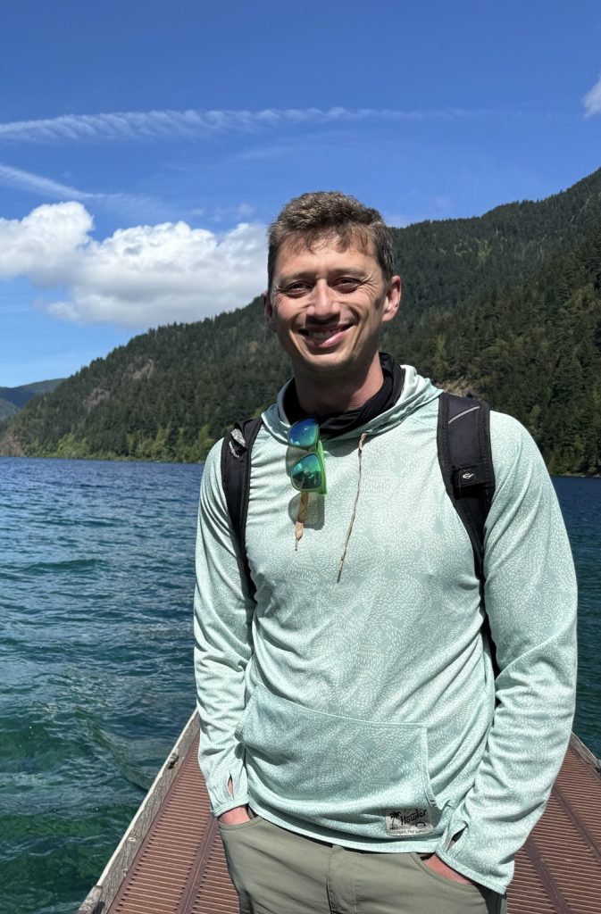 Scott Otto stands outdoors on a dock wearing a light green patterned hoodie, sunglasses hanging from the neck of his shirt, and a black backpack. Behind him is a scenic view of a lake surrounded by lush green forest and mountains under a bright blue sky with scattered clouds.