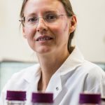 Portrait of a Natasha Kirienko in a white lab coat and glasses in a laboratory setting, with blurred sample bottles in the foreground.