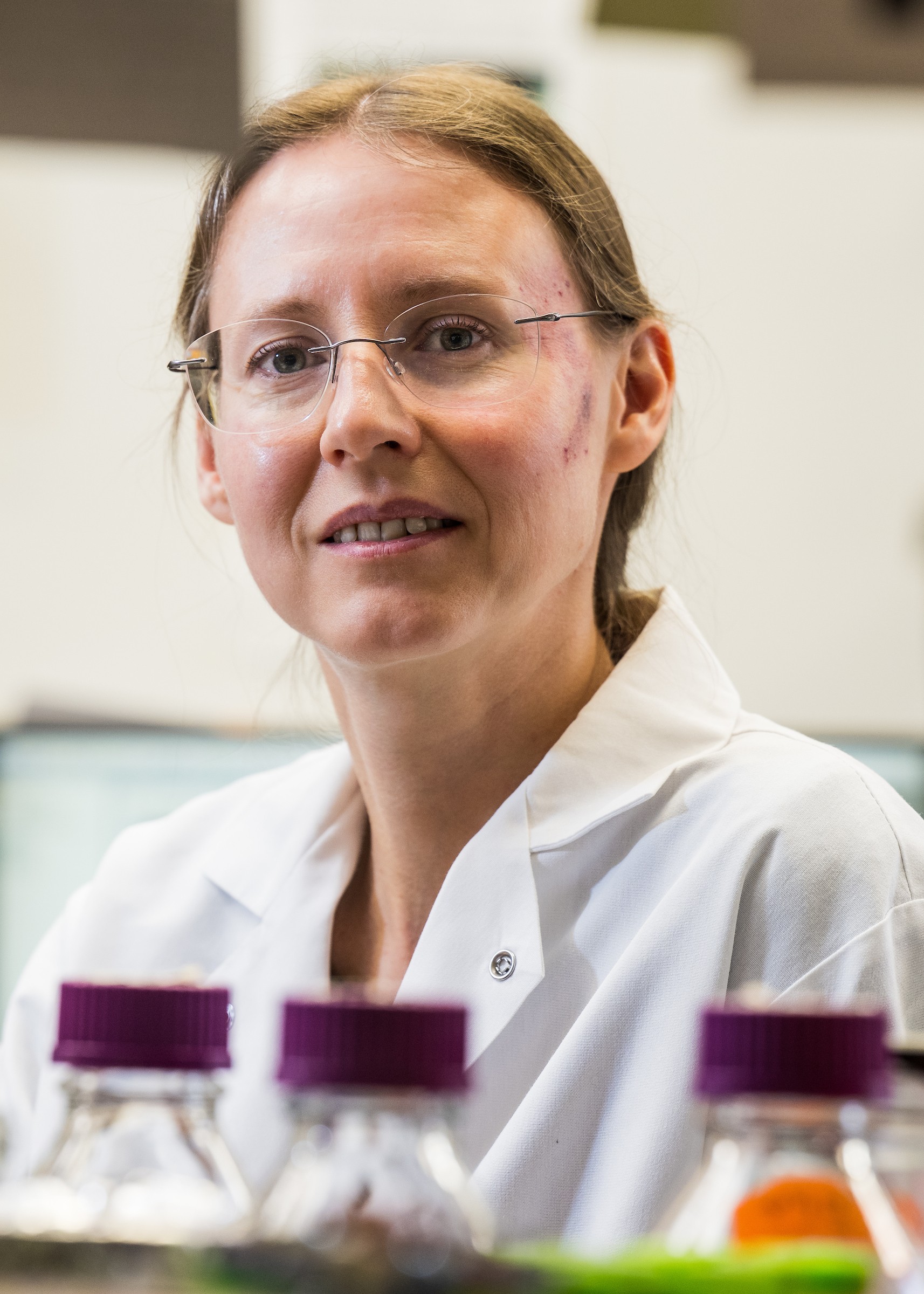 Portrait of a Natasha Kirienko in a white lab coat and glasses in a laboratory setting, with blurred sample bottles in the foreground.