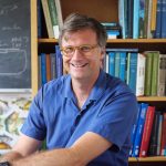 Dr. Carl Simpson smiles at the camera while he sits at a desk in an office with a bookshelf and blackboard in the background.