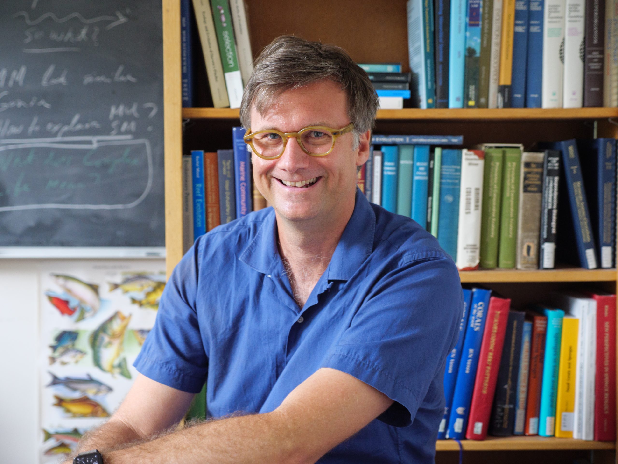 Dr. Carl Simpson smiles at the camera while he sits at a desk in an office with a bookshelf and blackboard in the background.