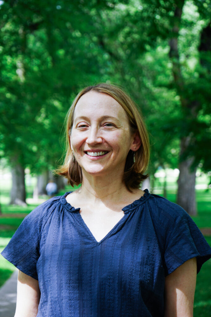 Dr. Jennifer Boyd stands amidst lush green sun dappled trees on the CSU campus while smiling warmly for her portrait. She wears a blue blouse with her hair down and cut short above the shoulders.