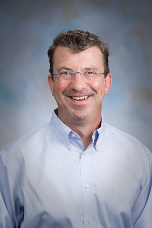 A formal headshot portrait of Dr. Joe von Fischer. He is smiling and wears glasses and a light blue button down shirt and sits in front of a blue-grey backdrop.