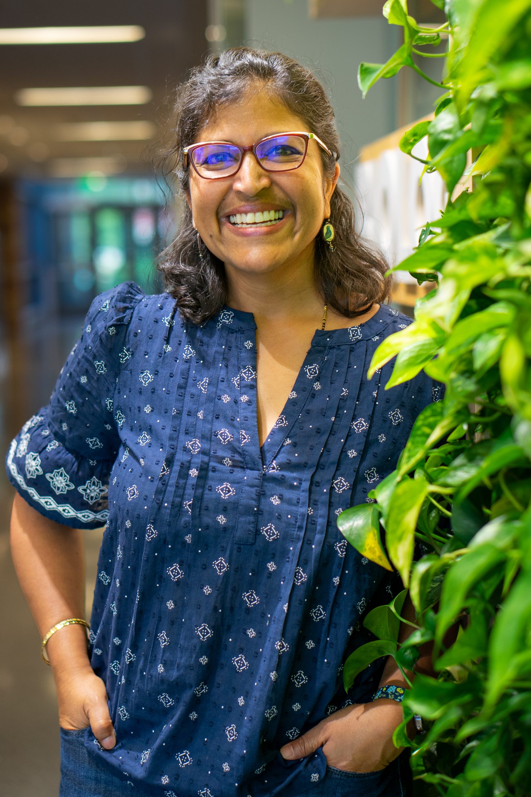 Portrait of Meena Balgopal, the current chair for CSU, Department of Biology. She wears glasses and a navy patterned blouse and smiles in the biology hallway beside the Living Wall of green plants.