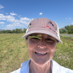 Ruth Hufbauer stands in an open grassy field with trees in the distance and a blue sky dotted with clouds above. She smiles at the camera while a praying mantis insect hangs out upside down on the underside of the brim of her baseball cap.