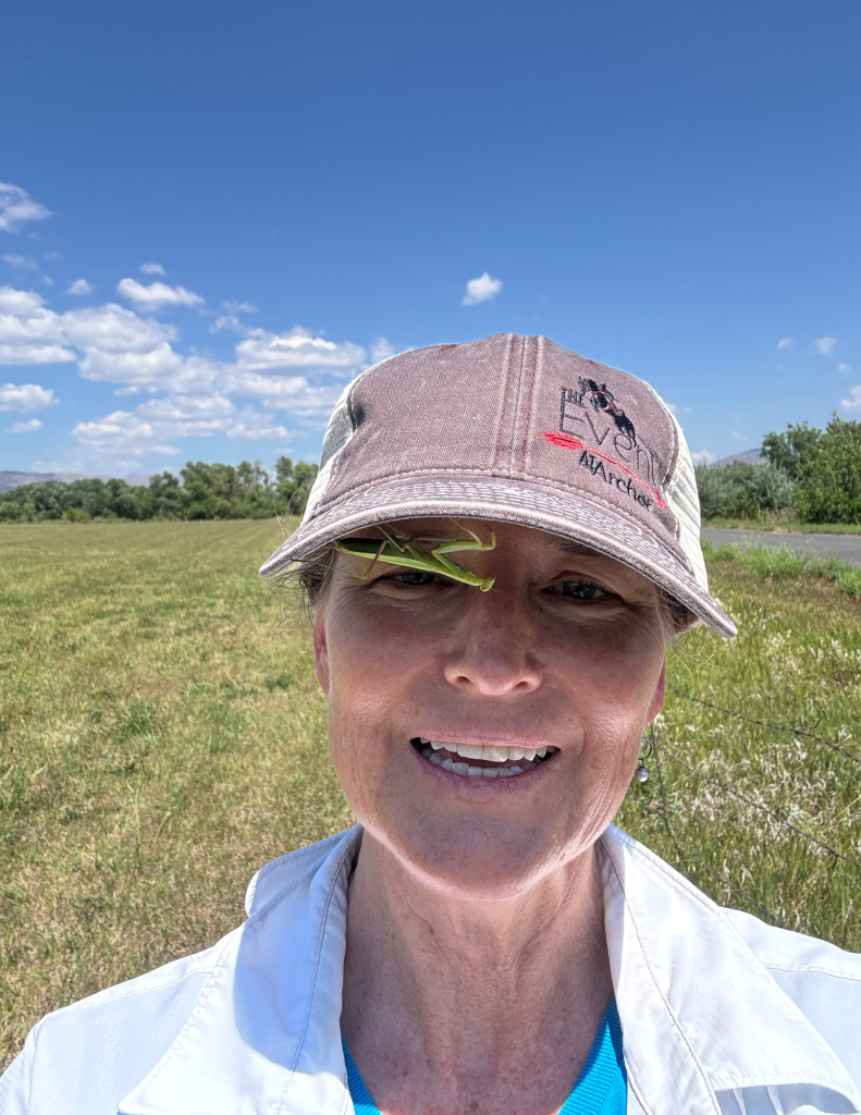 Ruth Hufbauer stands in an open grassy field with trees in the distance and a blue sky dotted with clouds above. She smiles at the camera while a praying mantis insect hangs out upside down on the underside of the brim of her baseball cap.